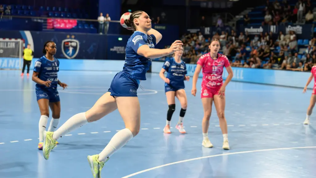 Une joueuse de handball en plein saut s'apprête à tirer au but dans une salle.