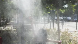 Une personne de dos, bras levés, jouant dans la brume rafraîchissante d'un parc urbain.