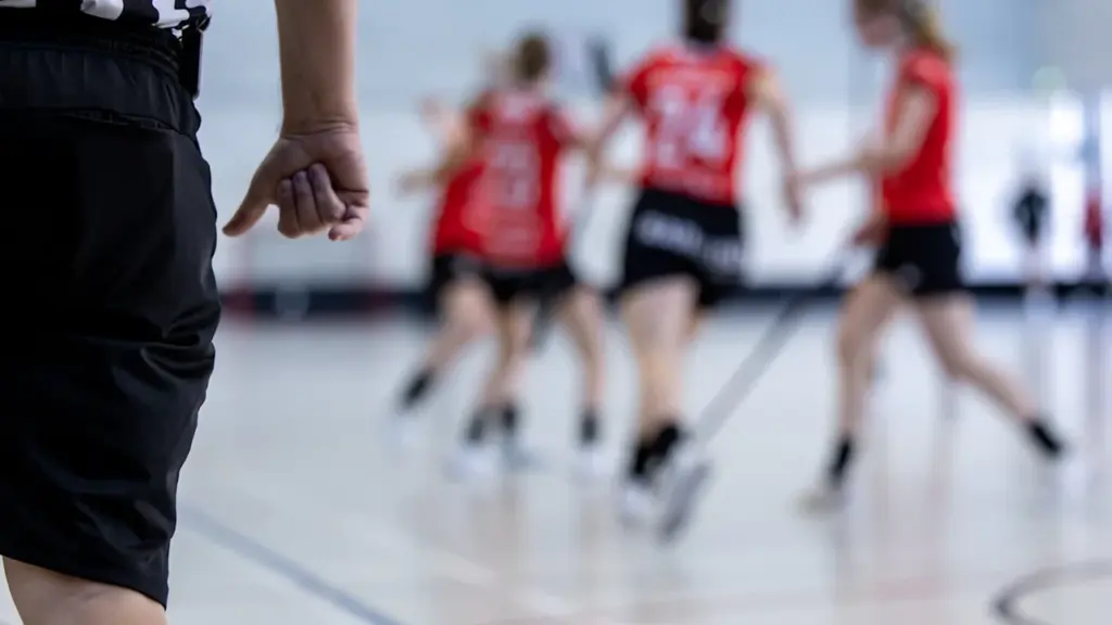 Une arbitre observe une équipe féminine évoluant sur un terrain de sport intérieur.