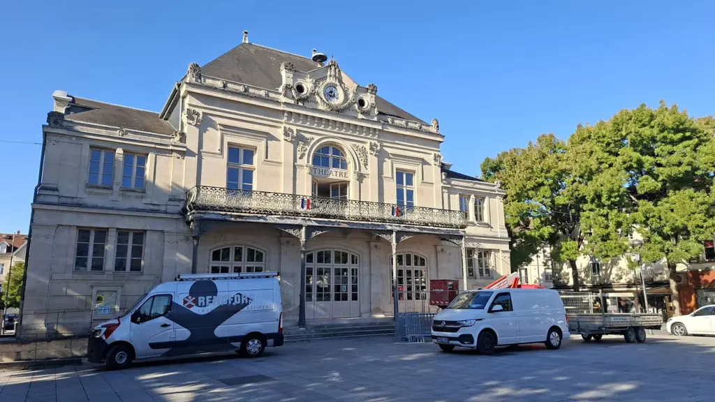 Façade d'un théâtre classique en pierre avec balcon devant lequel stationnent deux véhicules utilitaires.