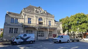 Façade d'un théâtre classique en pierre avec balcon devant lequel stationnent deux véhicules utilitaires.