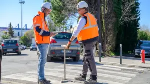 Deux ouvriers en gilets orange et casques blancs inspectent un passage piéton en extérieur.