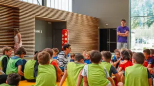 Un groupe d'enfants en chasubles colorées écoutent attentivement une femme assise dans un gymnase.