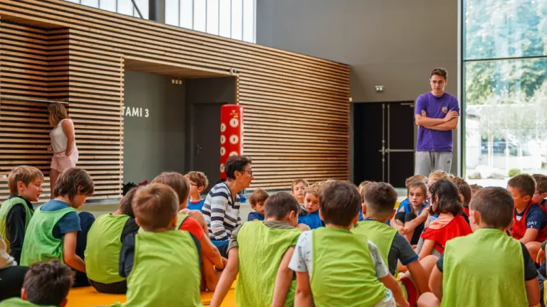 Un groupe d'enfants en chasubles colorées écoutent attentivement une femme assise dans un gymnase.