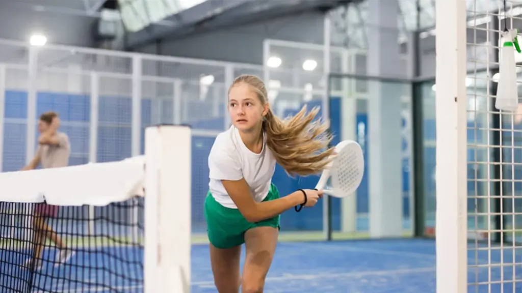 Une jeune fille aux cheveux blonds joue au padel sur un court intérieur bleu.