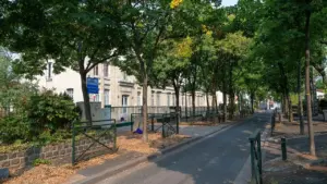 Une rue bordée d'arbres menant à une école primaire en pierre avec une grille métallique.